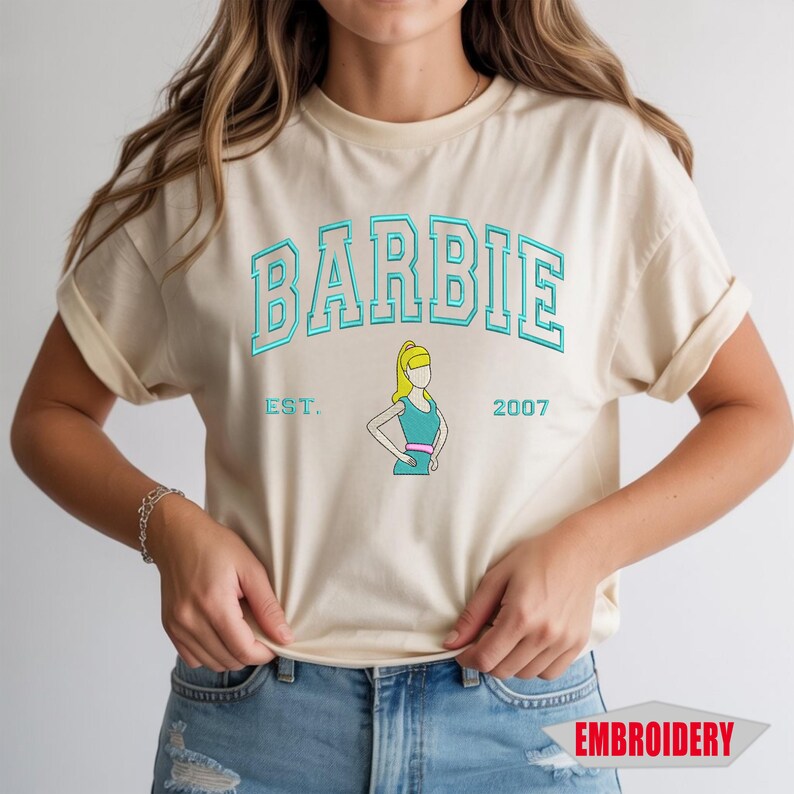 May include: A cream-colored short-sleeve t-shirt featuring the word BARBIE in a teal collegiate font. Below the text is a graphic of a blonde figure in a teal dress with a pink belt, flanked by the text EST. and 2007 in teal, with EMBROIDERY text.