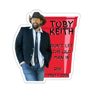 May include: A red plastic cup with the text "TOBY KEITH" and "DON'T LET THE OLD MAN IN" and "RIP 1961 - 2024". A man wearing a black jacket, a white shirt, a brown cowboy hat, and blue jeans is standing next to the cup.