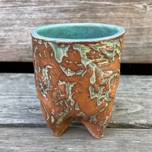 May include: A small, brown ceramic pot with a green glaze. The pot has three legs and a textured surface.