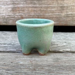 May include: A small, light green ceramic pot with three legs. The pot has a speckled glaze and a rounded shape.