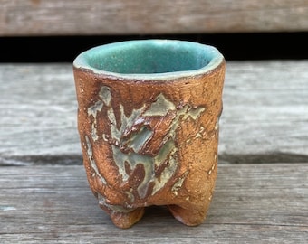 May include: A small, handmade ceramic cup with a textured, brown exterior and a teal interior. The cup has three small feet and a design of raised, leaf-like patterns. The cup is likely used for drinking.