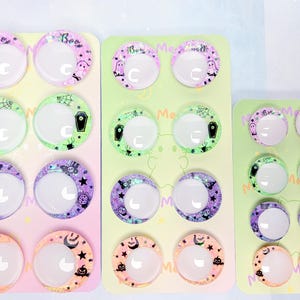 May include: Three rectangular display cards featuring pairs of circular cabochons with Halloween-themed designs. The prints include ghosts, coffins, spiderwebs, pumpkins, and stars in purple, green, and orange hues, each with a white crescent moon center.