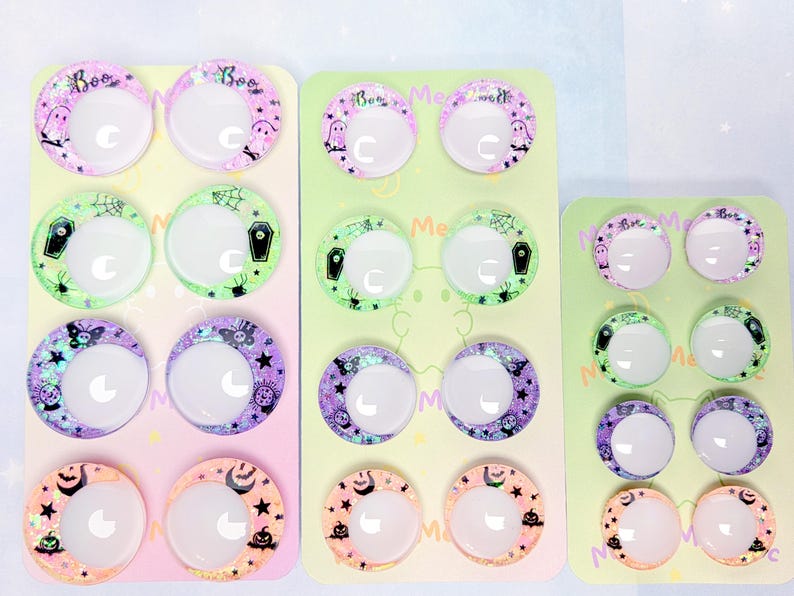 May include: Three rectangular display cards featuring pairs of circular cabochons with Halloween-themed designs. The prints include ghosts, coffins, spiderwebs, pumpkins, and stars in purple, green, and orange hues, each with a white crescent moon center.