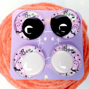 May include: Four circular cabochons on a light purple card. The top two have solid black centers, and the bottom two have solid white centers. Each features a pink glitter background with a ghost, a spider web, and the text Boo in black.