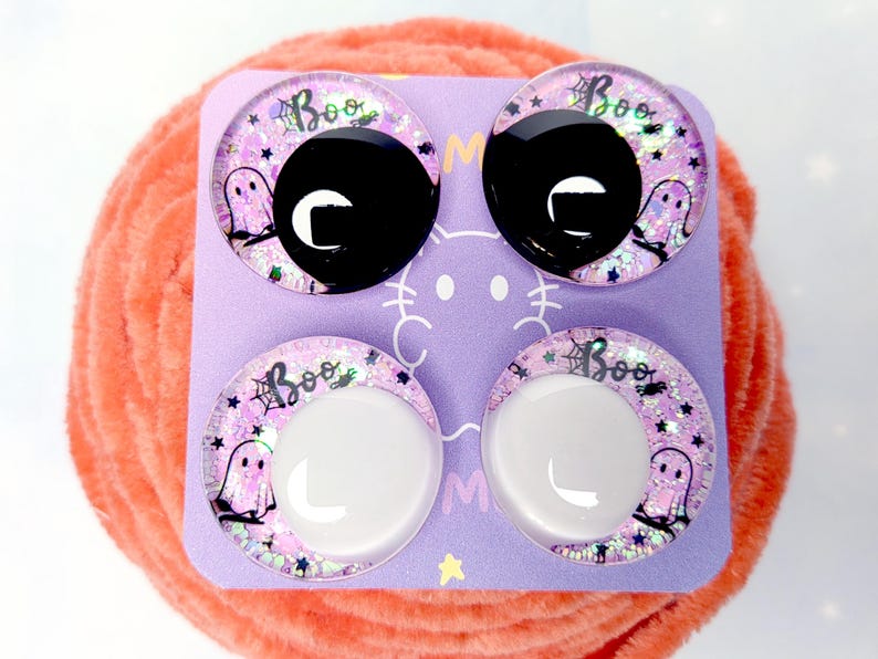 May include: Four circular cabochons on a light purple card. The top two have solid black centers, and the bottom two have solid white centers. Each features a pink glitter background with a ghost, a spider web, and the text Boo in black.