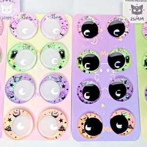 May include: A collection of circular 25mm resin cabochons featuring various Halloween-themed designs including ghosts, coffins, pumpkins, and stars. The cabochons are arranged on colorful display cards with the text MAKIN MEOWGIC and 25MM visible.