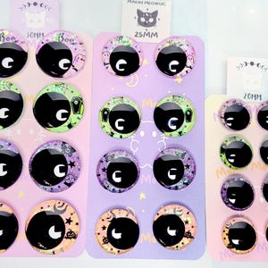 May include: Three display cards featuring pairs of round glass cabochon eyes with black pupils and colorful, patterned irises. The designs include ghosts, coffins, stars, and pumpkins in shades of purple, green, and orange. Labels indicate 25mm and 20mm sizes.
