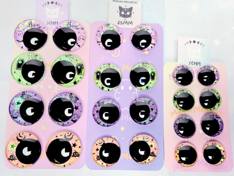 May include: Three display cards featuring pairs of round glass cabochon eyes with black pupils and colorful, patterned irises. The designs include ghosts, coffins, stars, and pumpkins in shades of purple, green, and orange. Labels indicate 25mm and 20mm sizes.