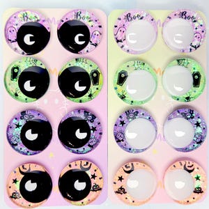 May include: Sixteen circular cabochons arranged on two rectangular display cards. The cabochons feature Halloween-themed designs including ghosts, coffins, spiders, and pumpkins with glittery backgrounds. Some have black pupils, while others are clear.