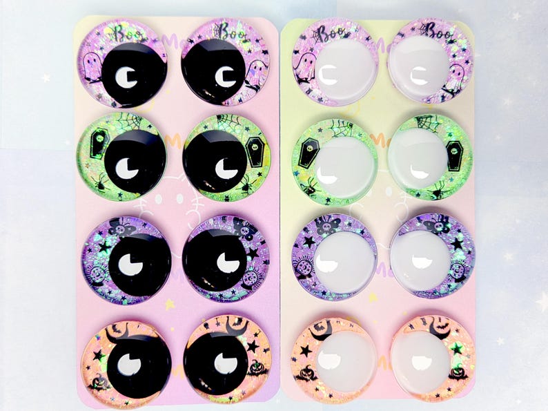 May include: Sixteen circular cabochons arranged on two rectangular display cards. The cabochons feature Halloween-themed designs including ghosts, coffins, spiders, and pumpkins with glittery backgrounds. Some have black pupils, while others are clear.