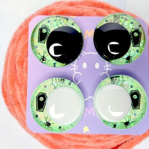 May include: Four circular cabochons arranged on a purple display card. The cabochons feature a light green glitter background with black spider web, spider, and coffin motifs. Two cabochons have large black pupils, while two have white pupils.