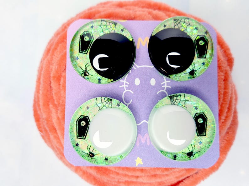 May include: Four circular cabochons arranged on a purple display card. The cabochons feature a light green glitter background with black spider web, spider, and coffin motifs. Two cabochons have large black pupils, while two have white pupils.