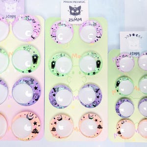 May include: Three vertical display cards holding pairs of circular cabochons with crescent moon cutouts. The cards are labeled 30mm, 25mm, and 20mm. The cabochons feature pastel-colored patterns including ghosts, coffins, spiders, bats, and pumpkins.