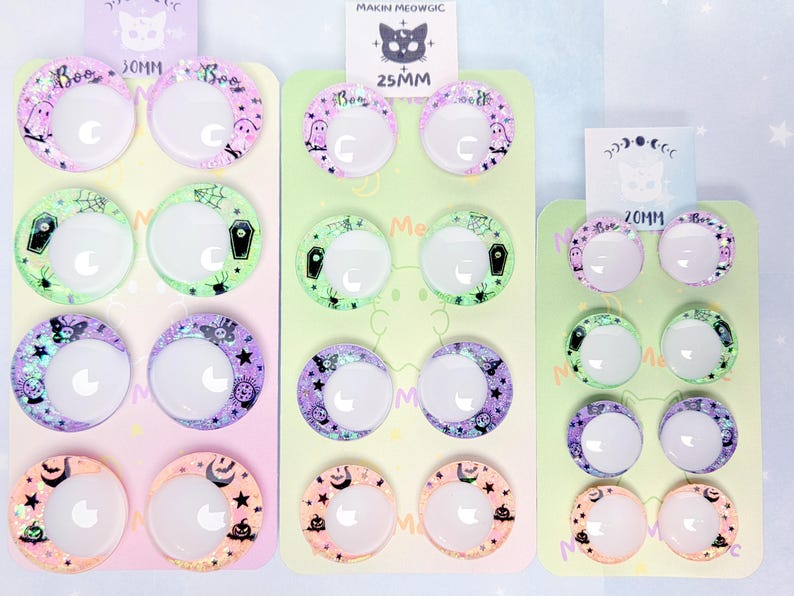 May include: Three vertical display cards holding pairs of circular cabochons with crescent moon cutouts. The cards are labeled 30mm, 25mm, and 20mm. The cabochons feature pastel-colored patterns including ghosts, coffins, spiders, bats, and pumpkins.