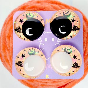 May include: Four circular cabochons arranged on a light purple display card. The top two feature black centers, while the bottom two have white centers. All four display a peach-colored background with black silhouettes of pumpkins, stars, and bats.