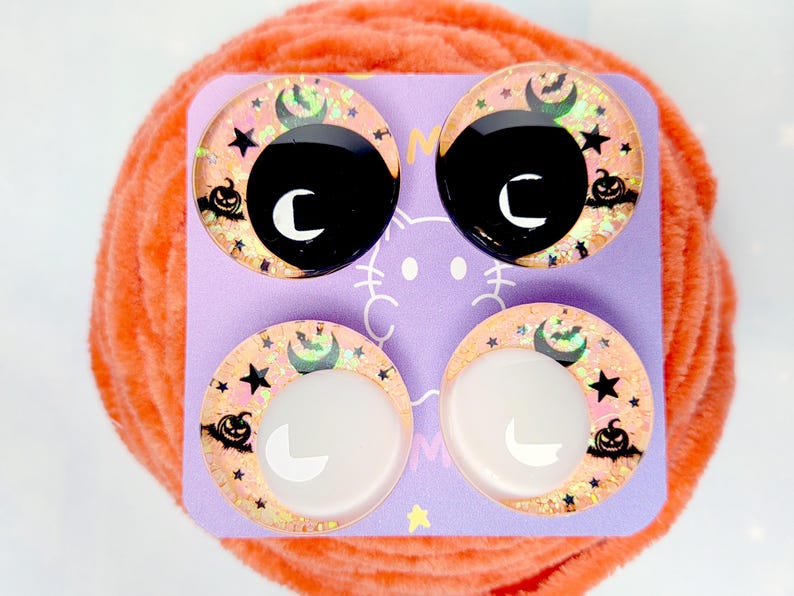 May include: Four circular cabochons arranged on a light purple display card. The top two feature black centers, while the bottom two have white centers. All four display a peach-colored background with black silhouettes of pumpkins, stars, and bats.