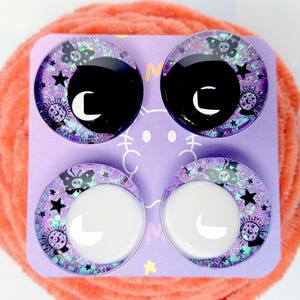 May include: Four circular cabochons arranged on a light purple card. The top two feature black centers with white crescent moons, while the bottom two have white centers with white crescent moons. All contain purple glitter, stars, butterflies, and crystal balls.
