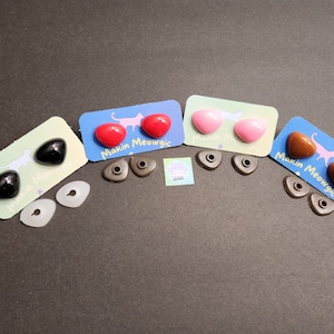 May include: Four pairs of plastic safety eyes for crafts, in black, red, pink, and brown. Each pair is packaged on a card with a cat illustration and the text "Makin Meowgic".