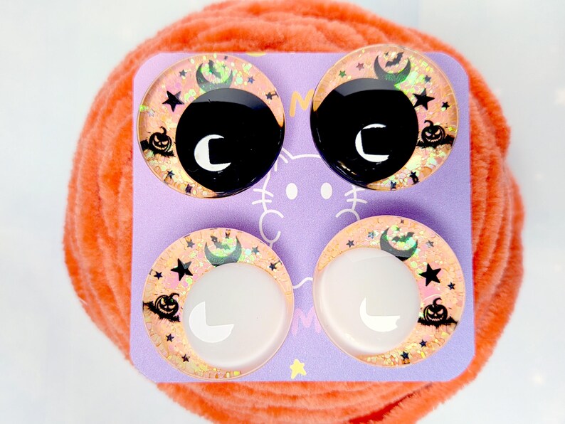 May include: Four circular cabochons arranged on a light purple display card. The top two feature black centers, while the bottom two have white centers. All four display a peach-colored background with black silhouettes of pumpkins, stars, and bats.