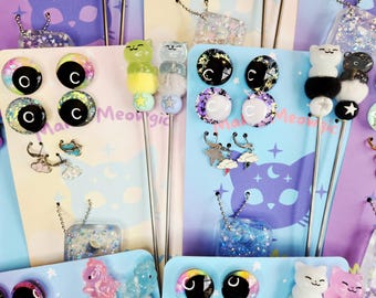 May include: A collection of metal hair sticks and jewelry sets displayed on branded cards. Each set features cat-themed resin charms, colorful pom-poms, star beads, and small cloud-shaped charms. The cards feature the text Make Meowgic.
