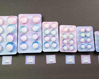 May include: Assortment of colorful, iridescent plastic eyes in various sizes, ranging from 16mm to 30mm. The eyes are arranged in rows on a white background with a paw print design. The text "Makin' Meowgic" is printed on one of the rows.