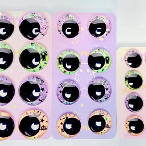 May include: A collection of round, domed glass cabochon eyes for crafting, featuring various Halloween-themed backgrounds including ghosts, coffins, crystal balls, and pumpkins. The eyes have large black pupils with white highlights, arranged on three display cards.