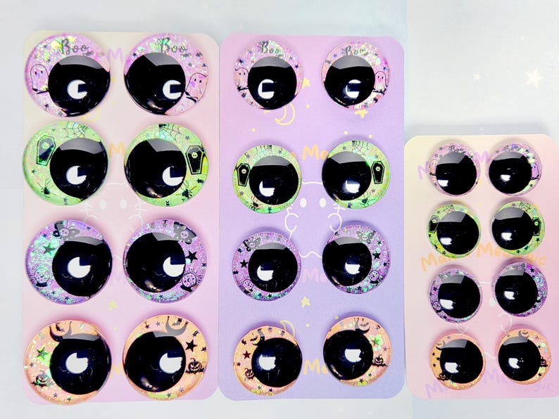 May include: A collection of round, domed glass cabochon eyes for crafting, featuring various Halloween-themed backgrounds including ghosts, coffins, crystal balls, and pumpkins. The eyes have large black pupils with white highlights, arranged on three display cards.