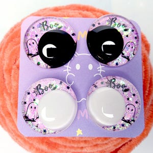 May include: Four circular cabochons on a light purple card. The top two have solid black centers, and the bottom two have solid white centers. Each features a pink glitter background with a ghost, a spider web, and the text Boo in black.
