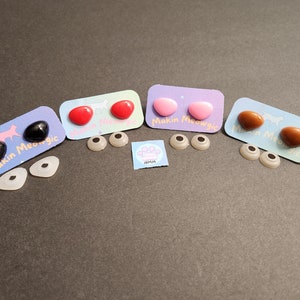 May include: Four sets of plastic safety eyes for crafts, with two eyes per set. The eyes are black, red, pink, and brown. Each set comes with a white card with the text "Makin Meowgic".