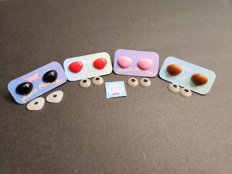 May include: Four sets of plastic safety eyes for crafts, with two eyes per set. The eyes are black, red, pink, and brown. Each set comes with a white card with the text "Makin Meowgic".