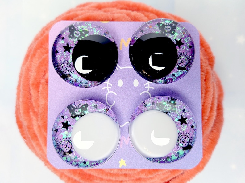 May include: Four circular cabochons arranged on a light purple card. The top two feature black centers with white crescent moons, while the bottom two have white centers with white crescent moons. All contain purple glitter, stars, butterflies, and crystal balls.