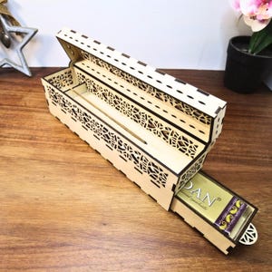 May include: A light-colored wooden incense box with intricate geometric cutouts. The box is open, revealing a compartment for incense sticks and a partially visible package of incense. The box is on a wooden surface.