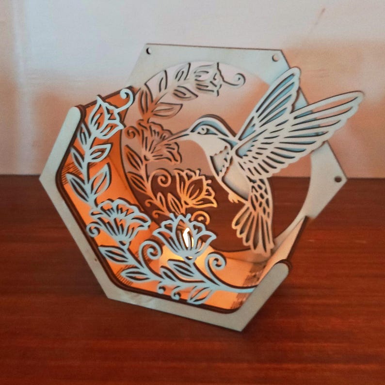 May include: A wooden, hexagonal hummingbird lantern. The lantern features a laser-cut hummingbird design with floral accents. The interior glows with an orange light, creating a warm ambiance. The outer frame is light-colored, with blue accents.