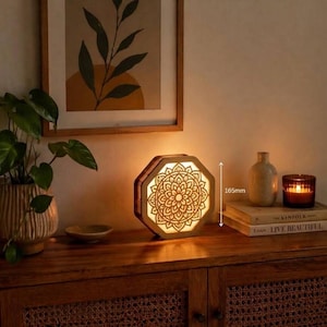 Può includere: Una lampada ottagonale in legno con un intricato motivo mandala tagliato al laser che emette una luce calda. La lampada è alta 165 mm, come indicato da una freccia di misurazione verticale, ed è posizionata su una superficie in legno.
