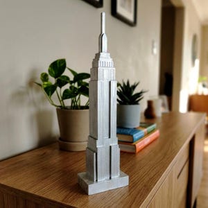 Può includere: Un modello tridimensionale color argento dell'Empire State Building. La struttura presenta un design a gradoni con una guglia centrale sulla sommità, poggiante su una base rettangolare. La superficie ha una finitura verticale testurizzata.