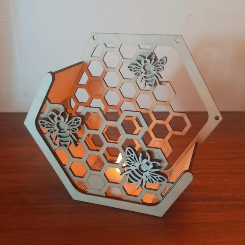 May include: A wooden, hexagonal candle holder with a honeycomb design and three decorative bee cutouts. The interior glows with warm light from a candle. The natural wood color contrasts with the orange glow, creating a cozy ambiance.