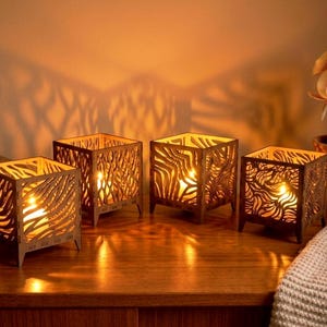 May include: Four square wooden candle holders with intricate cut-out designs, each with a lit candle inside. The candle holders are on a wooden surface, with a plant and a blanket in the background.