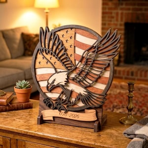Op de afbeelding: Een houten patriottische decoratie met een gedetailleerde lasergesneden Amerikaanse zeearend met uitgestrekte vleugels, tegen een achtergrond van de Amerikaanse vlag. Het ontwerp bevat de data "1776-2026" op een banner, met een gelaagde houten constructie.
