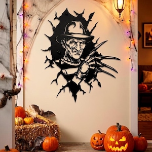 May include: A black metal wall decoration of a horror movie character breaking through a wall. The character is wearing a hat and has long, sharp claws. The scene is decorated with Halloween pumpkins, lights, and spiderwebs.