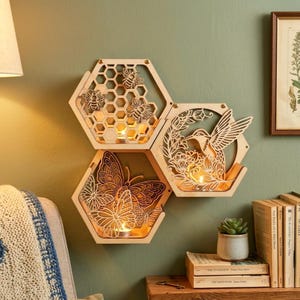 May include: Three wooden, hexagonal candle holders with detailed cut-out designs. One shows bees, another a butterfly, and the third a hummingbird. Each contains a lit candle, producing a warm glow. The holders are mounted on a green wall.