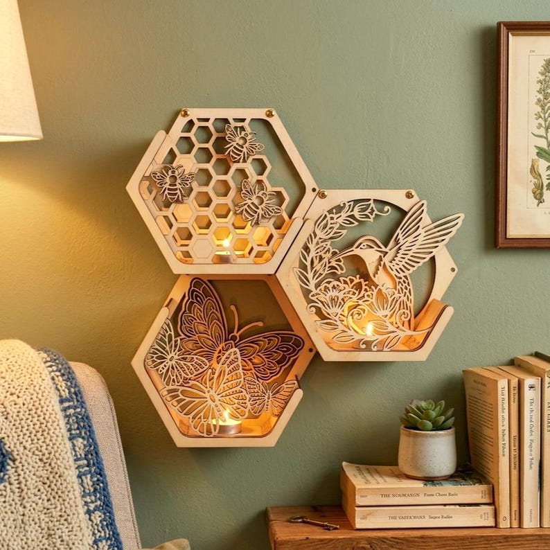 May include: Three wooden, honeycomb-shaped candle holders with intricate cut-out designs. One features bees, another a butterfly, and the third a hummingbird. Each holds a lit candle, creating a warm glow. The holders are mounted on a green wall.