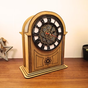 Peut inclure: Horloge de style cheminée en bois clair. Le cadran noir affiche des chiffres romains et un motif floral décoratif. Une fleur est gravée sur le devant. Les aiguilles rouges indiquent l'heure.
