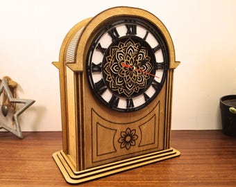 May include: Wooden mantel clock with a black clock face and Roman numerals. The clock features a decorative floral design in the center and a flower carved on the front. The clock is made of light brown wood.