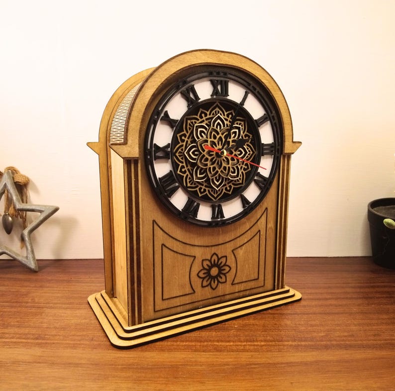 May include: Wooden mantel clock with a black clock face and Roman numerals. The clock features a decorative floral design in the center and a flower carved on the front. The clock is made of light brown wood.