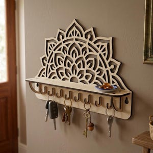 Può includere: Portachiavi in legno con un design mandala decorativo. Il portachiavi ha un ripiano e più ganci per le chiavi. Le chiavi sono appese ai ganci. Il portachiavi è montato su una parete di colore neutro.