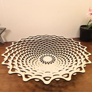 Op de afbeelding: Een decoratieve houten kom met een uniek, opengewerkt ontwerp. De kom heeft een herhalend patroon van gebogen vormen, waardoor een gelaagd, gestructureerd effect ontstaat. De natuurlijke houtkleur contrasteert met het donkere interieur, op een houten oppervlak.