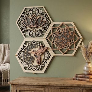 Puede incluir: Tres piezas de arte mural de madera con forma de hexágono. Cada pieza presenta un diseño en capas con una flor de loto, un ojo y un colibrí. La paleta de colores incluye tonos de madera natural, gris y rosa, creando una estética relajante.
