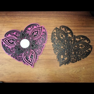 May include: Two heart-shaped decorative wooden pieces. One is layered with pink and black, featuring a floral design with a central circle. The other is a solid dark brown with a similar floral pattern. Both are on a wooden surface.