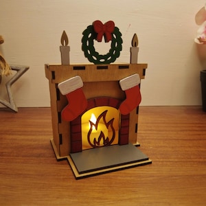 Puede incluir: Una decoración de chimenea navideña de madera con un efecto de llama brillante. La chimenea presenta calcetines rojos, una corona verde con un lazo rojo y dos acentos de velas. El diseño es festivo y de temporada.