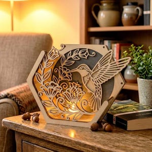 May include: A wooden, hexagonal light box featuring a hummingbird design. The hummingbird is intricately cut out, along with floral elements. The light box is placed on a wooden table, with acorns scattered around it. The light inside casts a warm glow.